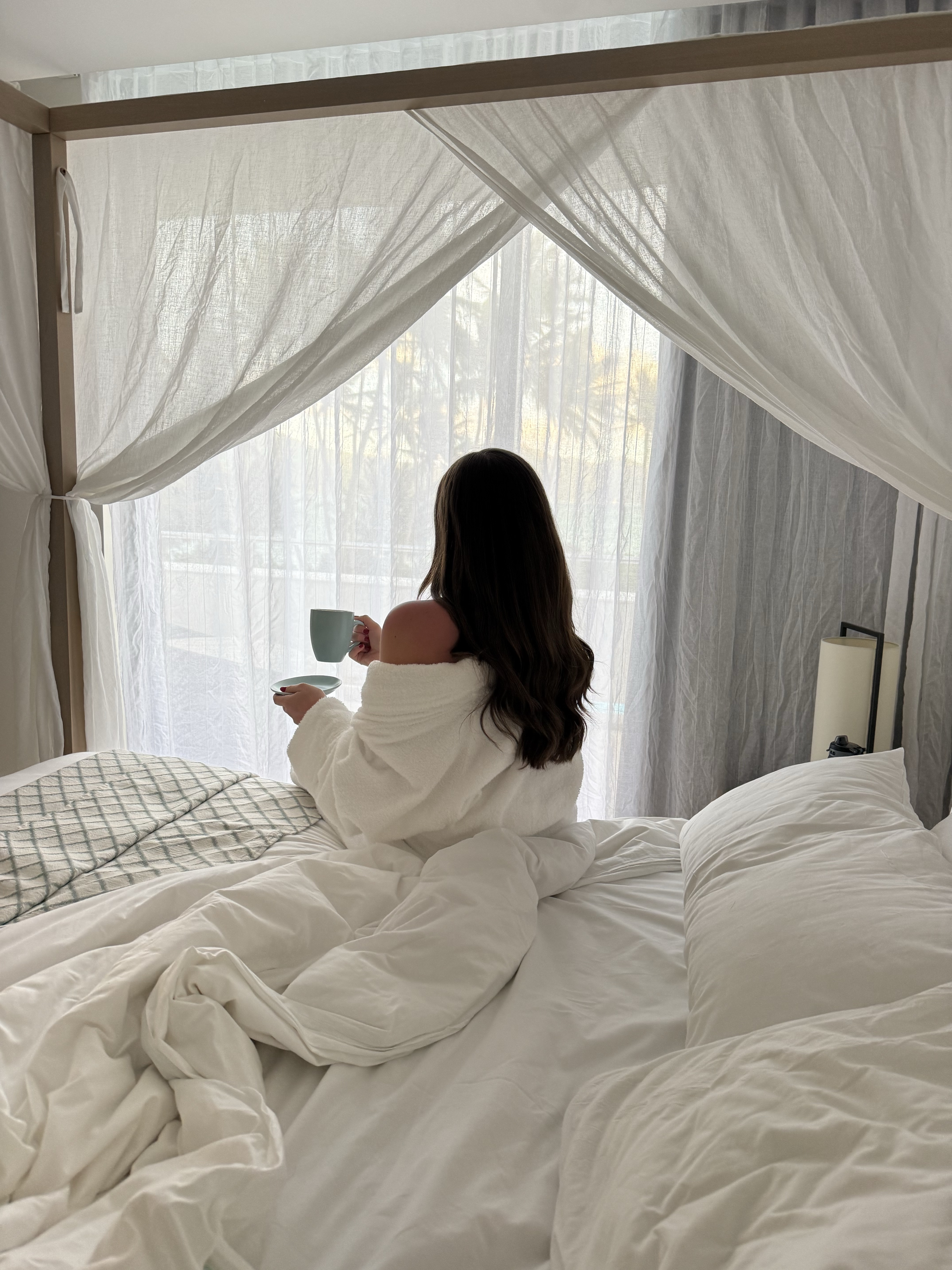 Woman sitting in bed with a cup by a sunlit window.